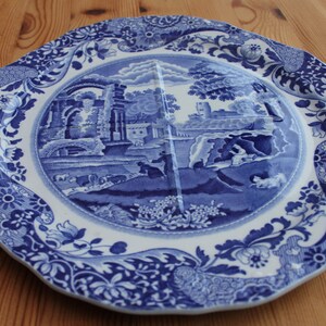 May include: Blue and white ceramic plate with a landscape scene in the center. The plate has a scalloped edge and floral border. The design features buildings, trees, and figures.