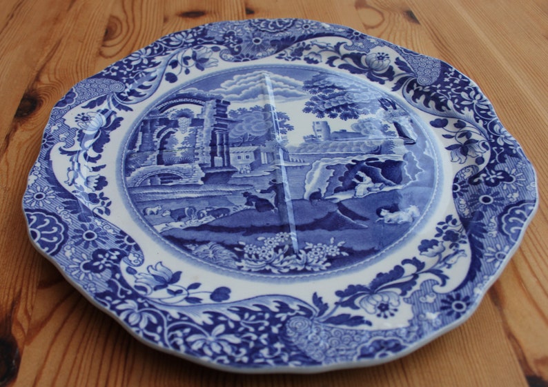 May include: Blue and white ceramic plate with a landscape scene in the center. The plate has a scalloped edge and floral border. The design features buildings, trees, and figures.