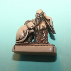 May include: A small, cream-colored carved figurine depicting a bearded figure with a long robe sitting beside a cat. The sculpture features intricate dark detailing within the grooves of the carving, set upon a smooth, rectangular base.
