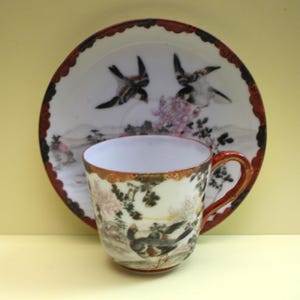 May include: A vintage teacup and saucer set. The white porcelain cup and saucer feature hand-painted images of birds, flowers, and landscapes. The cup has a red handle and trim, and the saucer has a red scalloped edge.