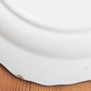 May include: A white ceramic plate with a scalloped edge. The plate has a slightly curved surface and a small imperfection on the edge. The plate is on a wooden surface.