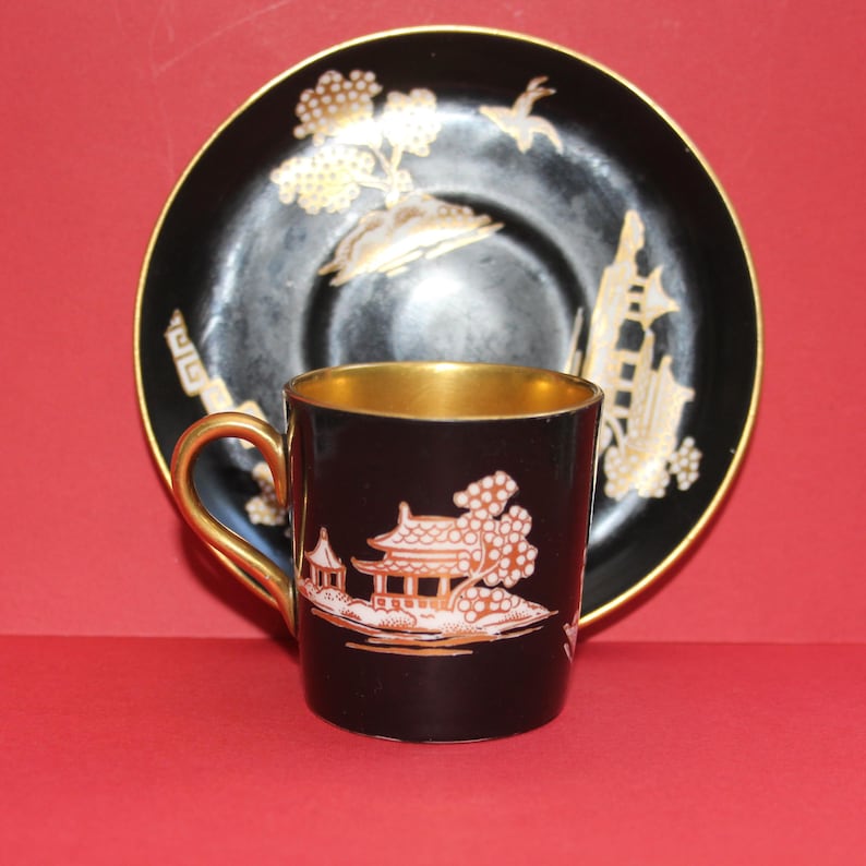 May include: A black teacup and saucer set with gold trim. The cup and saucer feature a gold and orange Asian-inspired design. The cup has a gold handle and interior. The saucer has a matching design. The set is on a red background.