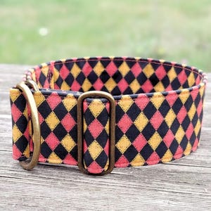May include: A wide dog collar with a repeating diamond pattern in black, coral, and gold. The collar has brass-colored metal hardware, including a D-ring and a rectangular buckle. The background is a blurred view of a wooden surface and greenery.