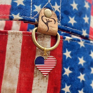 May include: A patriotic dog collar featuring a design of the American flag. The collar has a red, white, and blue pattern with stars and stripes. A heart-shaped charm with the American flag hangs from a gold ring.