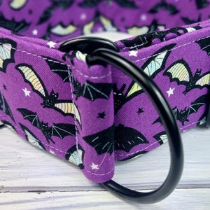 May include: A purple fabric collar featuring a repeating pattern of black bats with pale yellow wings and small white stars. The collar includes a sturdy black metal D-ring and a black metal slide adjuster, with visible white stitching along the edges.