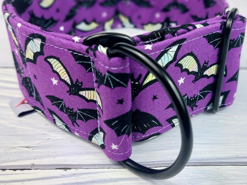 May include: A purple fabric collar featuring a repeating pattern of black bats with pale yellow wings and small white stars. The collar includes a sturdy black metal D-ring and a black metal slide adjuster, with visible white stitching along the edges.