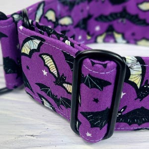 May include: A purple fabric collar featuring a repeating pattern of black bats with yellow and white wings, accented by small white stars. The collar includes black metal hardware, specifically a rectangular adjustment slider and a D-ring.