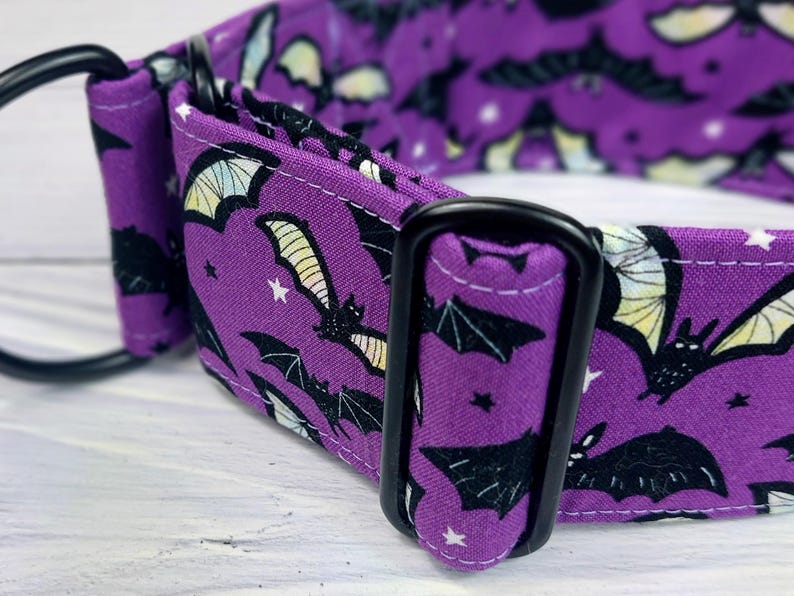 May include: A purple fabric collar featuring a repeating pattern of black bats with yellow and white wings, accented by small white stars. The collar includes black metal hardware, specifically a rectangular adjustment slider and a D-ring.