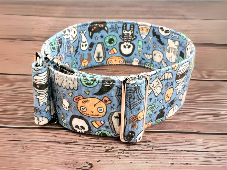 May include: A blue dog collar with a Halloween-themed print featuring ghosts, skeletons, mummies, and other spooky characters. The collar has a silver buckle.