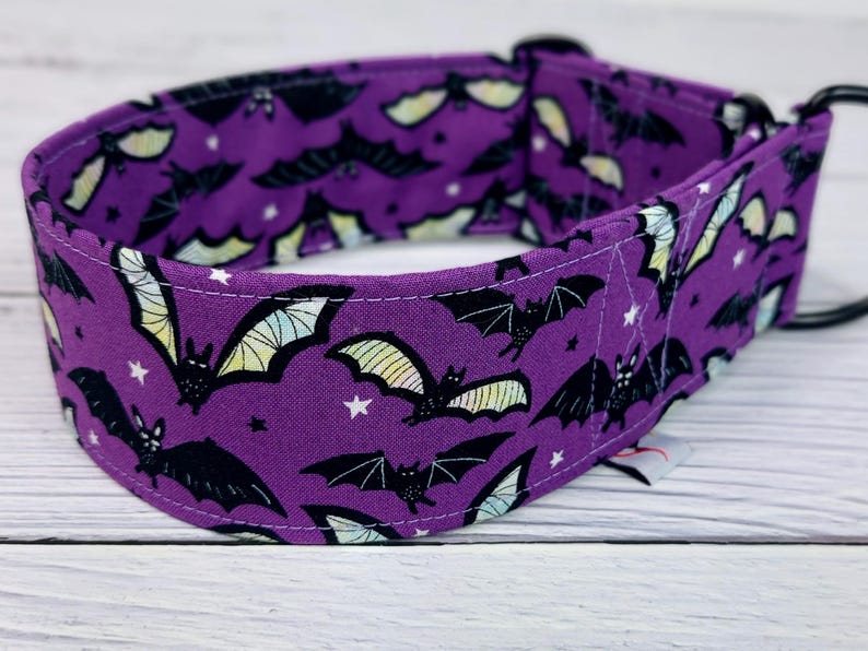 May include: A purple fabric pet collar featuring a repeating pattern of black bats with pale yellow and green wings, accented by small white stars. The collar includes a black metal D-ring and a black plastic buckle closure for secure fastening.