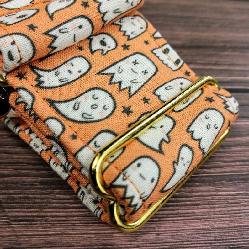 May include: A close-up of a bright orange fabric dog collar with a gold metal buckle. The fabric has a repeating pattern of white ghosts with black eyes and mouths. The ghosts are on a peach background with small black stars.