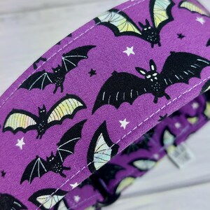May include: A purple fabric collar featuring a repeating pattern of black bats with light yellow and white wings, interspersed with small white stars. The collar has visible white stitching along the edges and a black plastic buckle component.