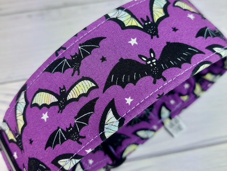 May include: A purple fabric collar featuring a repeating pattern of black bats with light yellow and white wings, interspersed with small white stars. The collar has visible white stitching along the edges and a black plastic buckle component.