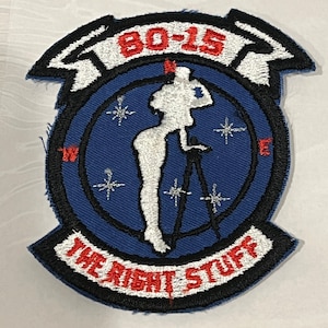 Pu&ograve; includere: Una toppa ricamata blu e bianca con un bordo nero. La toppa presenta una silhouette bianca di una donna in piedi su un treppiede con una macchina fotografica. Il testo "80-15" &egrave; in rosso e il testo "The Right Stuff" &egrave; in bianco.