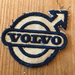 May include: A white and blue embroidered patch with the Volvo logo. The logo features a circle with a smaller circle inside, and the word "VOLVO" written in a blue, bold font.