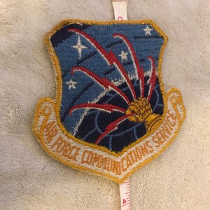 May include: A blue and gold embroidered patch with the text "Air Force Communications Service" and a stylized image of a globe with red and white lightning bolts radiating outward.