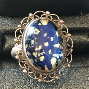 May include: Oval-shaped silver ring with a dark blue stone speckled with light green and yellow flecks. The stone is set in a decorative filigree frame with small beaded accents. The ring is a vintage or antique style.