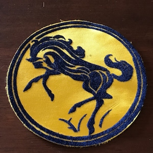 May include: A round patch featuring a navy blue horse silhouette on a yellow background. The horse is in a dynamic pose, with its legs extended. The patch has a navy blue border.