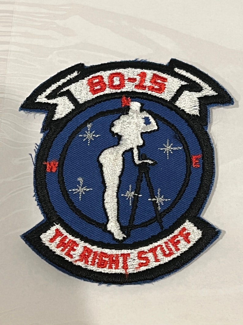 Pu&ograve; includere: Una toppa ricamata blu e bianca con un bordo nero. La toppa presenta una silhouette bianca di una donna in piedi su un treppiede con una macchina fotografica. Il testo "80-15" &egrave; in rosso e il testo "The Right Stuff" &egrave; in bianco.