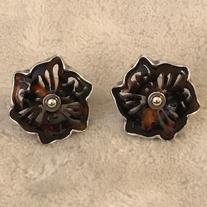 May include: A pair of brown and silver flower-shaped earrings. The earrings are made of a dark brown material with a silver metal frame. The center of each earring has a small silver bead.