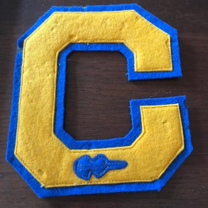 May include: A felt letter C with a blue border and a blue outline of a small figure inside the letter. The letter is yellow.