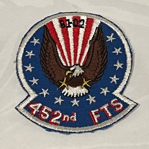 May include: A blue and white embroidered patch with a bald eagle in flight, a red, white, and blue American flag, and the text "452nd FTS".