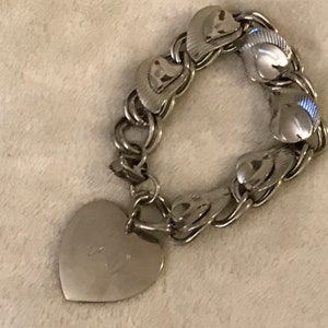 May include: A silver charm bracelet with a heart-shaped charm. The bracelet is made of a chain with heart-shaped links. The charm has a small inscription on it.