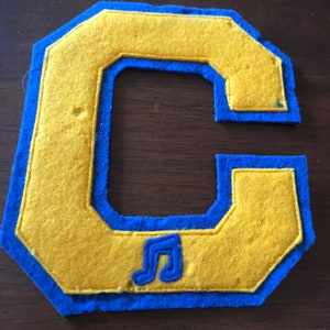 May include: A felt letter C in yellow with a blue border. The letter has a musical note design in the center.