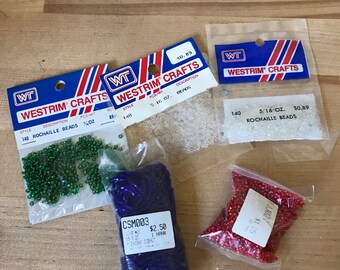 May include: Three bags of beads, one with green beads, one with blue beads, and one with red beads. The bags are labeled with the brand name "Westrim Crafts" and the price and weight of the beads. The bags are on a wooden surface.
