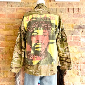 May include: Camouflage jacket with a graphic of a person's face and the words "are you experienced" in a yellow and red design. The jacket has a button-up front and long sleeves. The background is a brick wall.