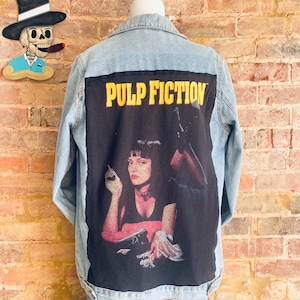 Pode incluir: Uma jaqueta de ganga azul claro com um remendo preto nas costas. O remendo apresenta uma cena do filme Pulp Fiction com o texto "PULP FICTION" em letras amarelas.