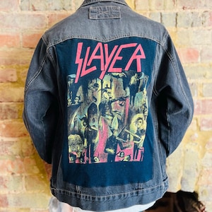 Può includere: Giacca di jeans grigio scuro con pannello posteriore nero con il nome della band "SLAYER" in lettere rosa. Il pannello posteriore mostra anche una grafica colorata della band.