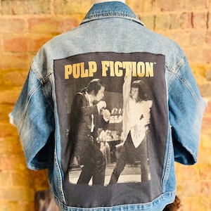 May include: Light blue denim jacket with a large black and white graphic of a scene from the movie "Pulp Fiction" on the back. The words "PULP FICTION" are printed in gold letters above the graphic.