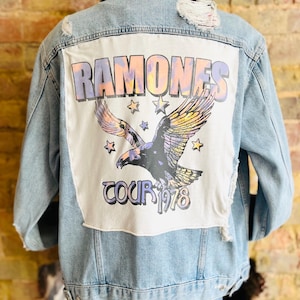 May include: Light blue denim jacket with a distressed back. The back features a white patch with the words "RAMONES TOUR 1978" and an eagle graphic in shades of purple, yellow, and black. The jacket has a classic collar and button closures.