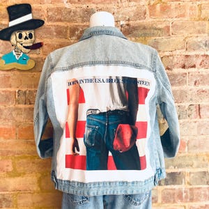 May include: Light blue denim jacket with a graphic on the back featuring a person in jeans and a red hat, with the text "BORN IN THE USA/BRUCE SPRINGSTEEN". The jacket is displayed on a mannequin.