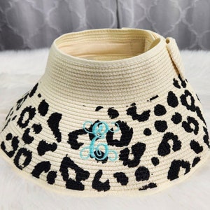 Puede incluir: Un sombrero de paja blanco con un dise&ntilde;o de estampado de leopardo negro. El sombrero tiene un ala ancha y un monograma bordado turquesa.