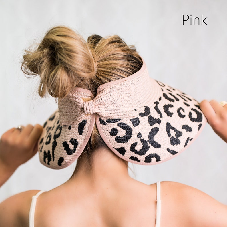 Puede incluir: Una visera de sol de tejido tejido con estampado de leopardo rosa y negro y detalle de lazo. La visera tiene un ala ancha.