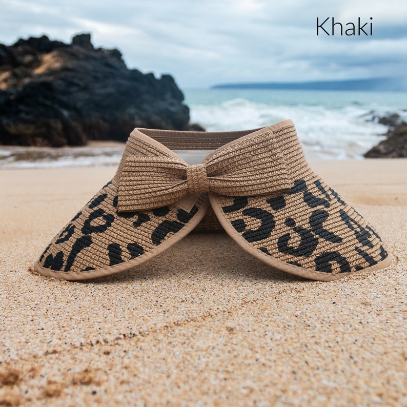 Puede incluir: Una visera de sol de color caqui con estampado de leopardo negro y marr&oacute;n y un gran lazo. La visera est&aacute; tumbada en una playa de arena.