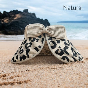 Puede incluir: Una visera de sol de paja beige y negra con estampado de leopardo y un gran lazo en la parte superior. La visera est&aacute; hecha de paja y tiene un ala ancha. La visera est&aacute; sentada en una playa de arena.