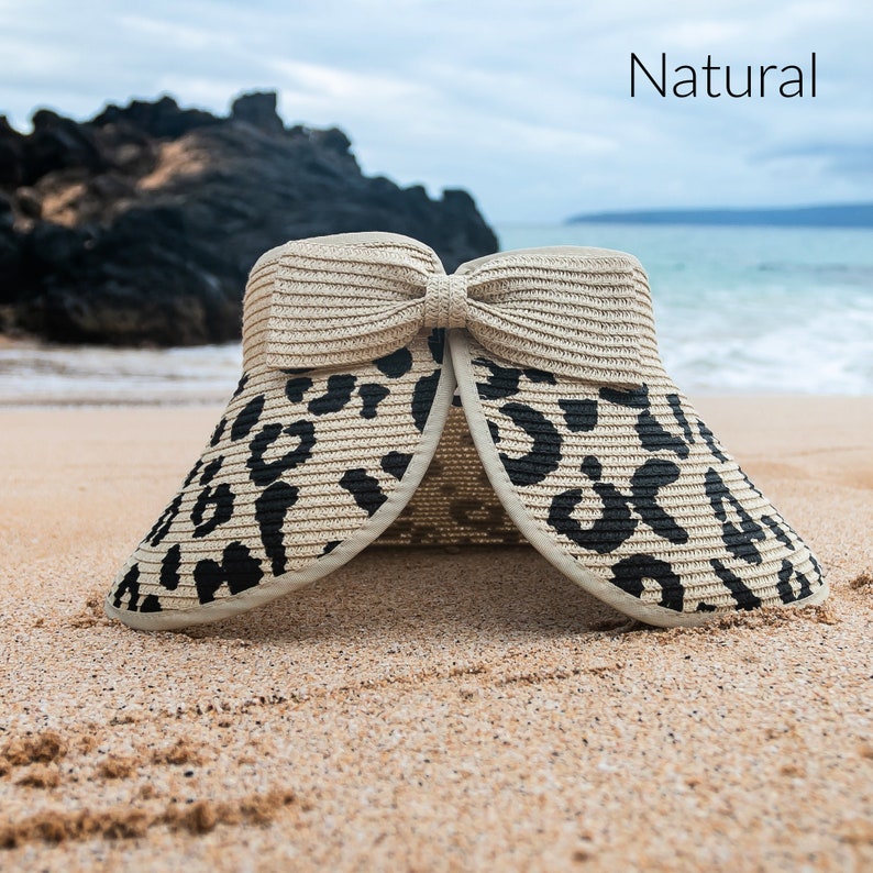 Puede incluir: Una visera de sol de paja beige y negra con estampado de leopardo y un gran lazo en la parte superior. La visera est&aacute; hecha de paja y tiene un ala ancha. La visera est&aacute; sentada en una playa de arena.