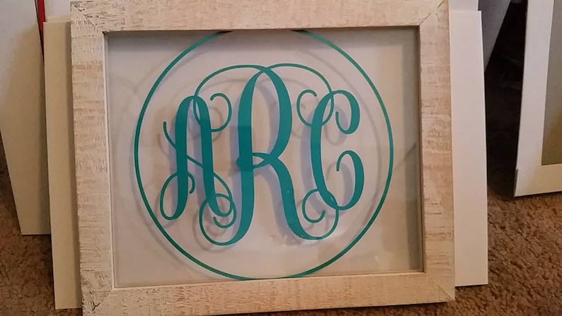 May include: A white wooden frame with a clear glass insert. The glass insert features a teal monogram of the letters "ARC" in a circle.