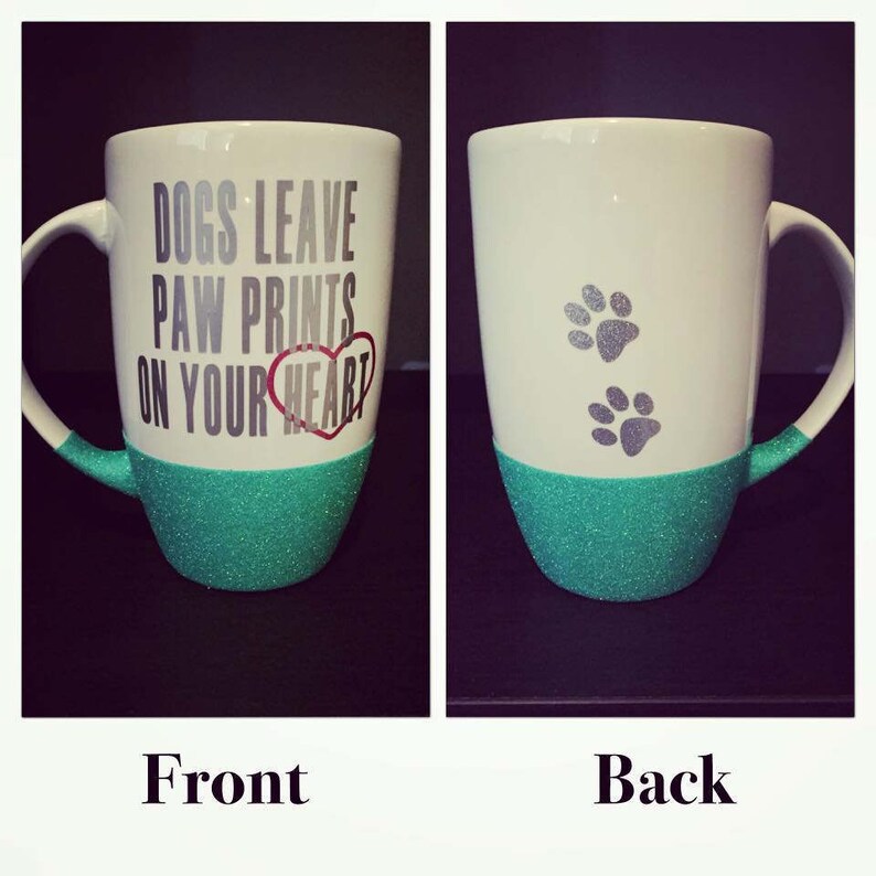 May include: White ceramic mug with a teal glitter base. The front of the mug has the text "Dogs leave paw prints on your heart" in black lettering with a red heart. The back of the mug has two silver paw prints.