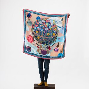 May include: A colorful silk scarf with a hot air balloon design. The balloon is filled with flowers and has a blue and gold pattern. The scarf has a pink, blue, and white border.