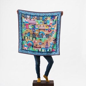 May include: A person is holding up a blue and brown patterned scarf with a cityscape design. The scarf has a blue border with a white and gold pattern.