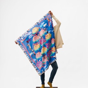 May include: A blue scarf with a colorful floral pattern and whimsical characters. The scarf is being held by a person wearing jeans and a beige sweater.