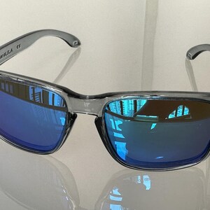 K&ouml;nnte beinhalten: Graue Sonnenbrille mit blau verspiegelten Gl&auml;sern. Die Sonnenbrille hat einen rechteckigen Rahmen und einen gebogenen B&uuml;gel. Die Gl&auml;ser spiegeln die Umgebung wider. Der Text "U.S.A." ist auf dem linken B&uuml;gel sichtbar.