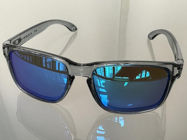 K&ouml;nnte beinhalten: Graue Sonnenbrille mit blau verspiegelten Gl&auml;sern. Die Sonnenbrille hat einen rechteckigen Rahmen und einen gebogenen B&uuml;gel. Die Gl&auml;ser spiegeln die Umgebung wider. Der Text "U.S.A." ist auf dem linken B&uuml;gel sichtbar.