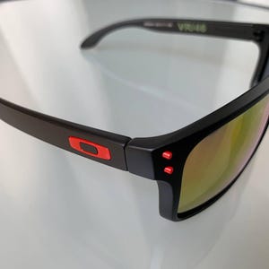 May include: Black sunglasses with a rectangular frame and mirrored lenses. The sunglasses have red accents on the temples and the Oakley logo. The lenses reflect a golden hue.