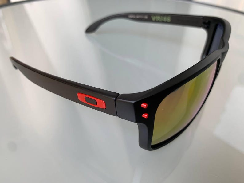 May include: Black sunglasses with a rectangular frame and mirrored lenses. The sunglasses have red accents on the temples and the Oakley logo. The lenses reflect a golden hue.