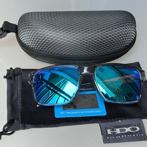 K&ouml;nnte beinhalten: Quadratische Sonnenbrille mit blau verspiegelten Gl&auml;sern, einem schwarzen Etui und einem schwarzen Beutel. Die Sonnenbrille hat einen transparenten Rahmen. Enth&auml;lt eine kleine blaue Karte und ein Etikett mit der Marke "HDO".